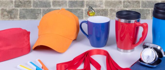 Promotional products neatly arranged on a gray desk, including a variety of mugs, lanyards, a ballcap, a notebook, and pens.