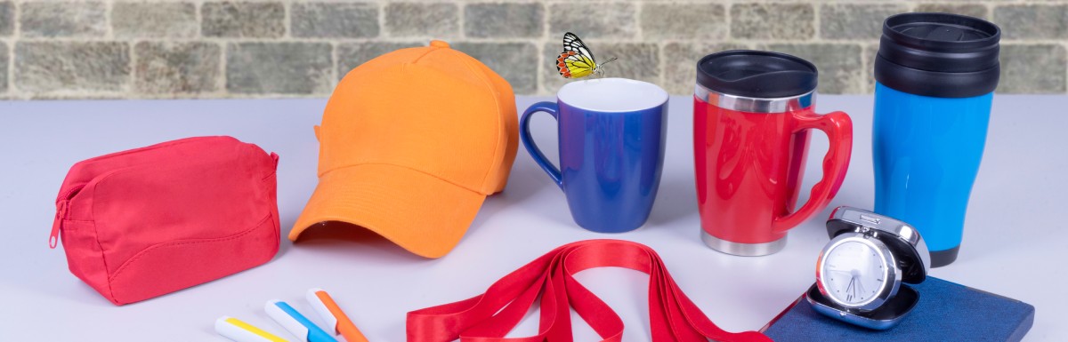 Promotional products neatly arranged on a gray desk, including a variety of mugs, lanyards, a ballcap, a notebook, and pens.