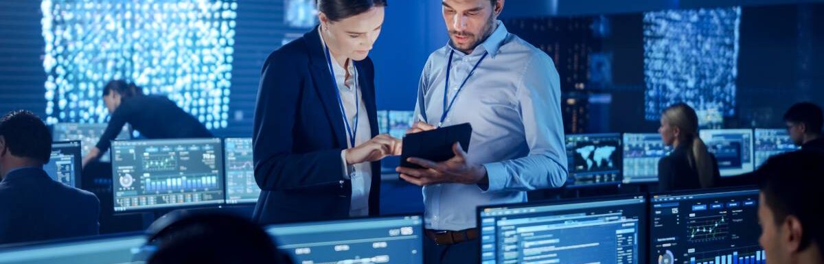 Two employees look at data on a tablet while standing in a room full of people reading data on monitors.