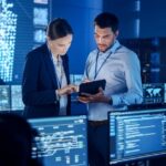 Two employees look at data on a tablet while standing in a room full of people reading data on monitors.