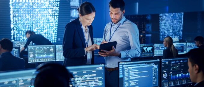 Two employees look at data on a tablet while standing in a room full of people reading data on monitors.