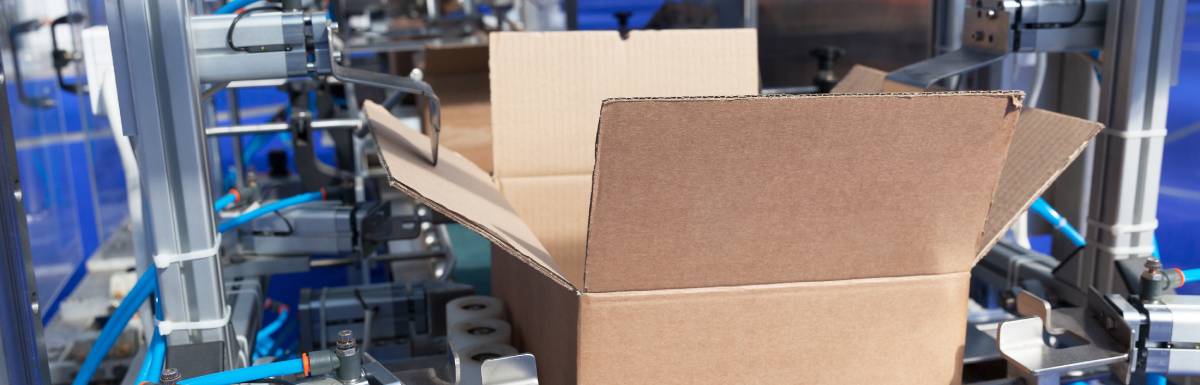 A cardboard box on the conveyor belt of a packaging machine, its outer flaps held open by two mechanical arms.