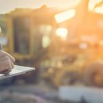 An engineer stands in front of a construction site while using a tablet to manage the project's logistics.