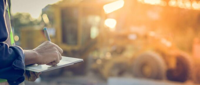 An engineer stands in front of a construction site while using a tablet to manage the project's logistics.