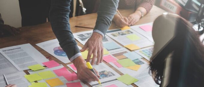 Men and women surround a table covered in various business-related sheets that include Venn diagrams, reports, and images.