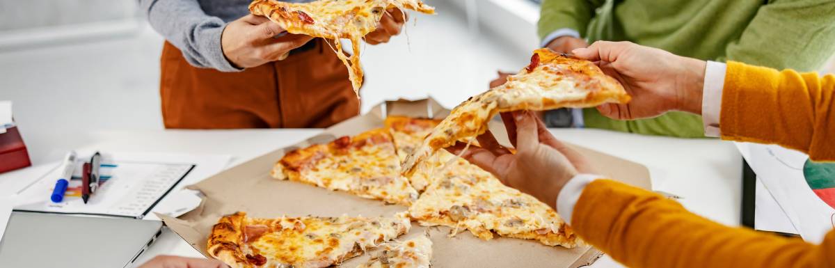 A small group of people are gathered around a desk to enjoy an office pizza party. Everybody is grabbing a slice.