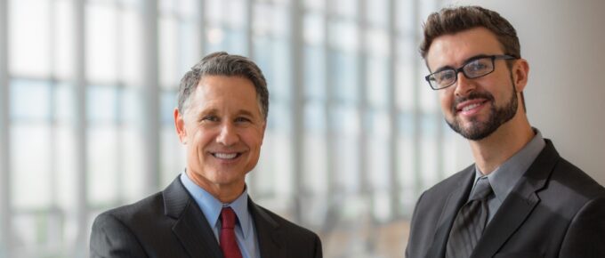 Two businessmen standing in an office building, side by side, and shaking hands. Both wear business suits.