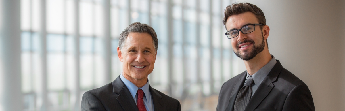 Two businessmen standing in an office building, side by side, and shaking hands. Both wear business suits.