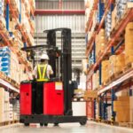 A worker, wearing a hard hat and a safety vest, uses a fork lift truck to move wooden pallets in a warehouse.