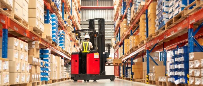 A worker, wearing a hard hat and a safety vest, uses a fork lift truck to move wooden pallets in a warehouse.