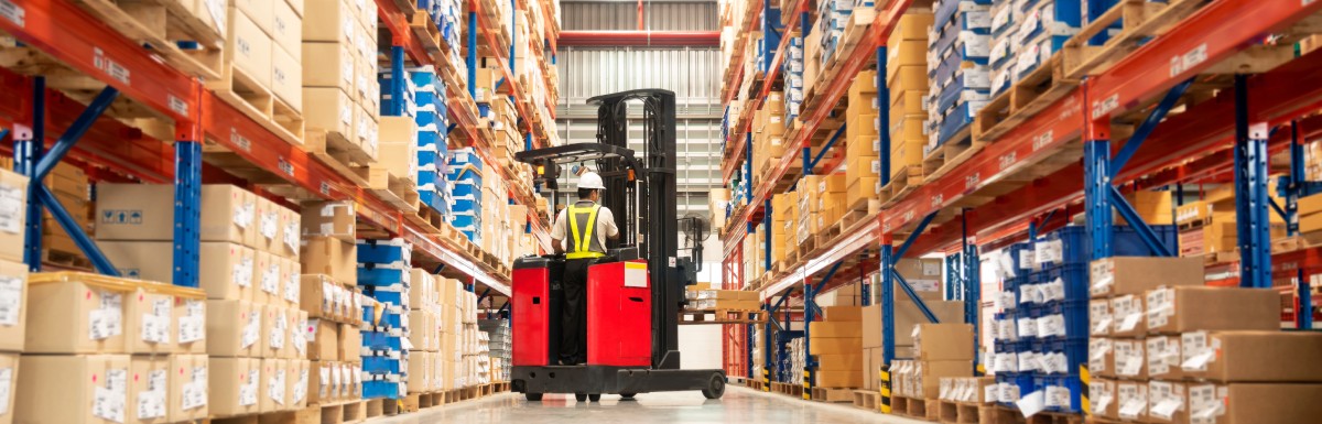 A worker, wearing a hard hat and a safety vest, uses a fork lift truck to move wooden pallets in a warehouse.