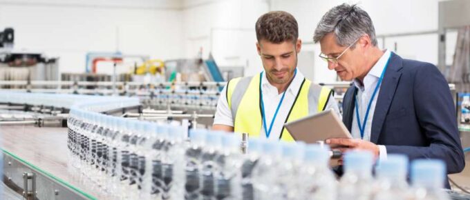 Two men standing on a manufacturing floor in front of a conveyor belt. One is in a suit and the other is in a safety vest.