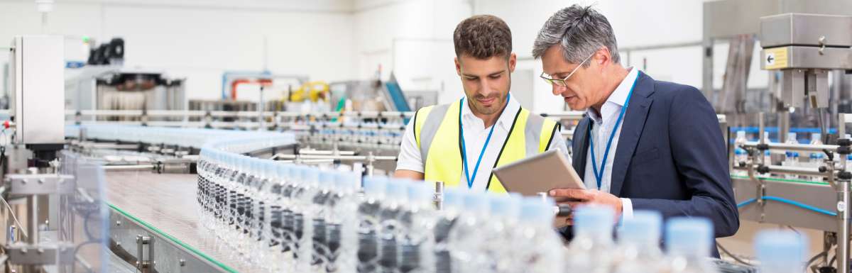 Two men standing on a manufacturing floor in front of a conveyor belt. One is in a suit and the other is in a safety vest.