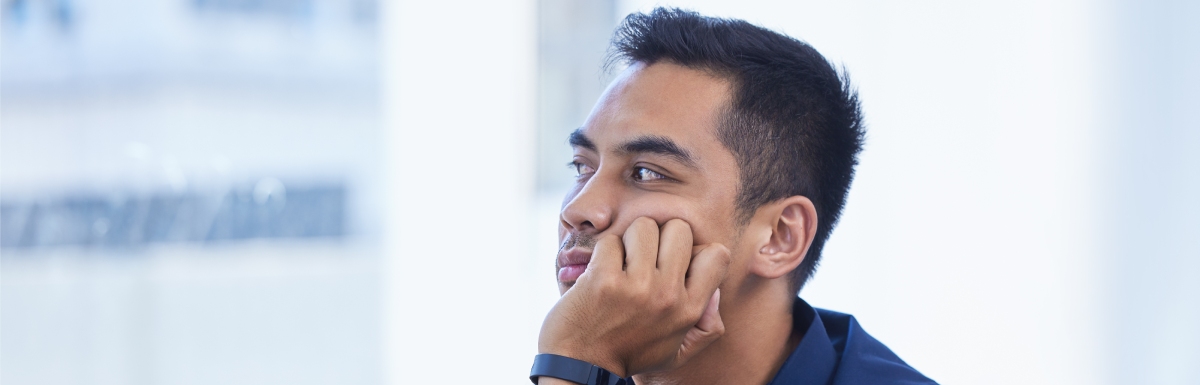 A man in a blue button-down shirt sits at an office desk, looking into the distance with his head on his cheek.