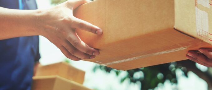 A man in a delivery uniform hands a sealed cardboard box to a customer in the middle of the day while holding another box.