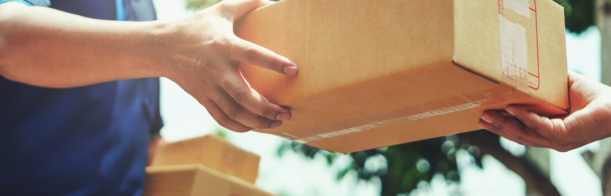 A man in a delivery uniform hands a sealed cardboard box to a customer in the middle of the day while holding another box.