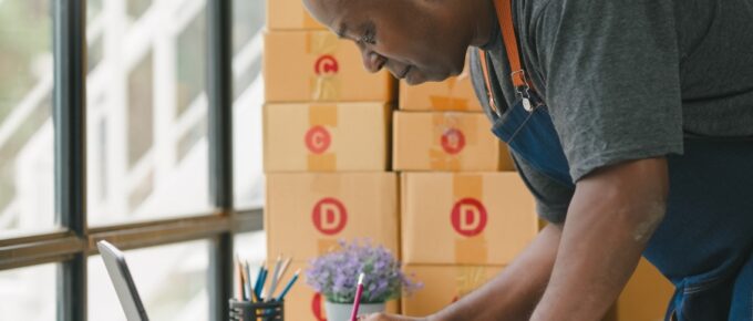 A Black man in an apron leaning over his work desk to fill out a form on a clipboard. There are packages stacked behind him.