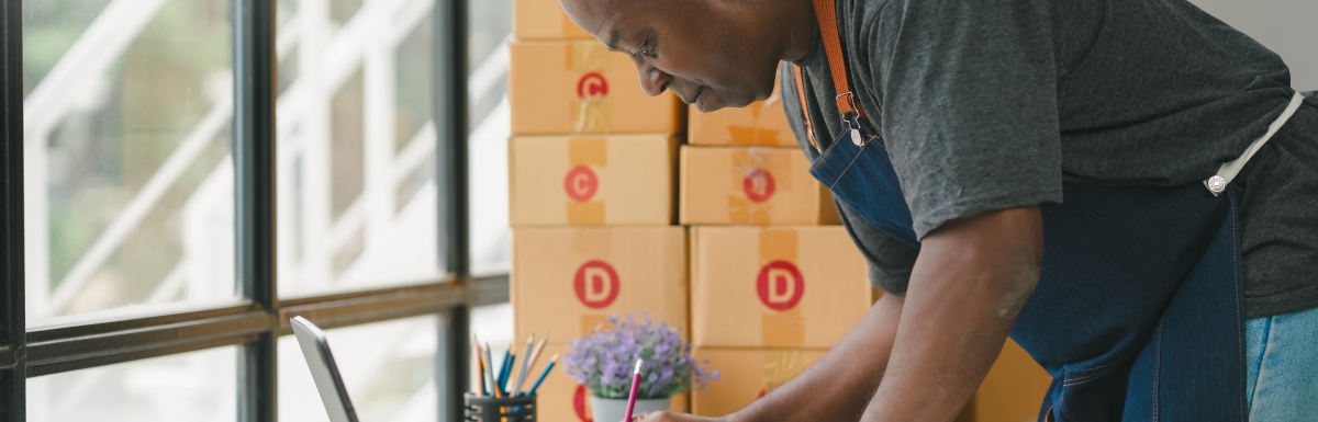 A Black man in an apron leaning over his work desk to fill out a form on a clipboard. There are packages stacked behind him.