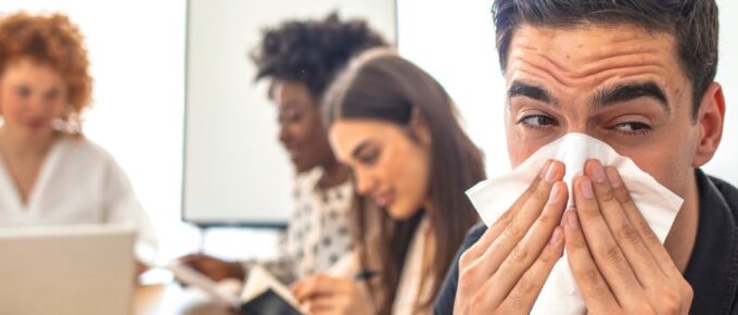 A man with a furrowed brow sneezing into a tissue toward the camera with three coworkers blurred in the background.