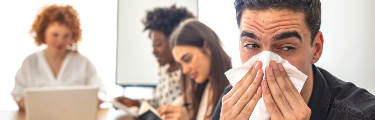 A man with a furrowed brow sneezing into a tissue toward the camera with three coworkers blurred in the background.