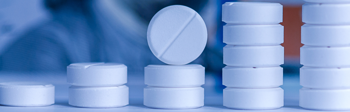 Five stacks of pills stand in front of a background of a $100 bill featuring the face of Benjamin Franklin.