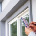 An inspector looks at the outside of a house. He checks something off his list using a blue clipboard.