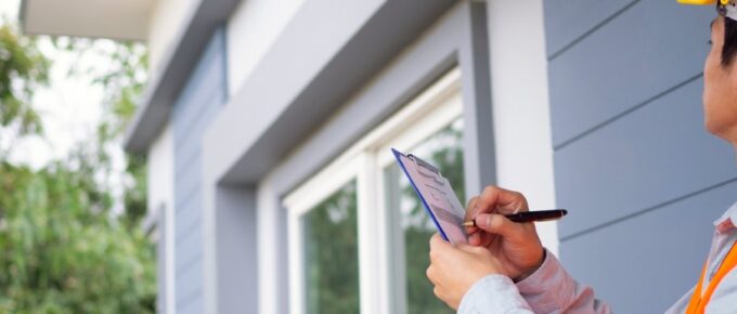 An inspector looks at the outside of a house. He checks something off his list using a blue clipboard.