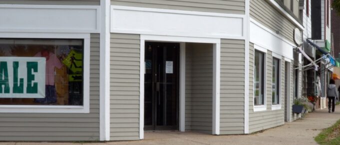 A relatively generic looking gray strip mall storefront is on the corner of an intersection. A sign in the window reads "SALE".