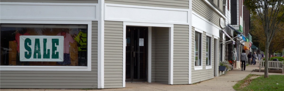 A relatively generic looking gray strip mall storefront is on the corner of an intersection. A sign in the window reads "SALE".