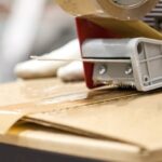 Gloved hands using a tape dispensing tool to seal the top of a cardboard box with clear packaging tape.