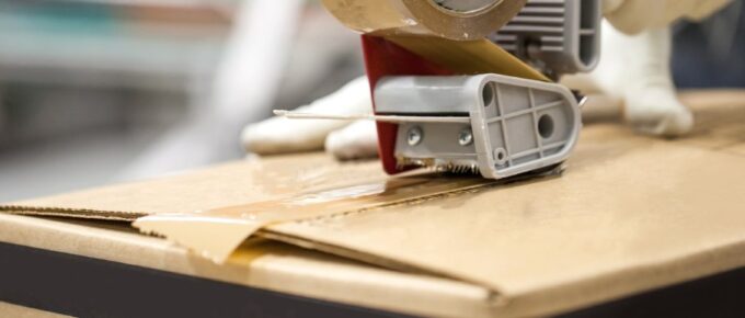 Gloved hands using a tape dispensing tool to seal the top of a cardboard box with clear packaging tape.