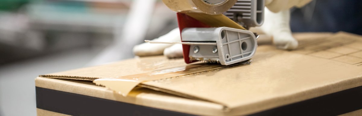 Gloved hands using a tape dispensing tool to seal the top of a cardboard box with clear packaging tape.