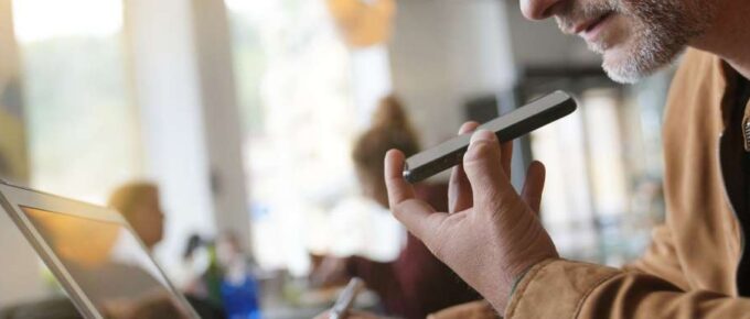 A middle-aged man sitting in a busy coffee shop and speaking into his cellphone while viewing his open laptop.