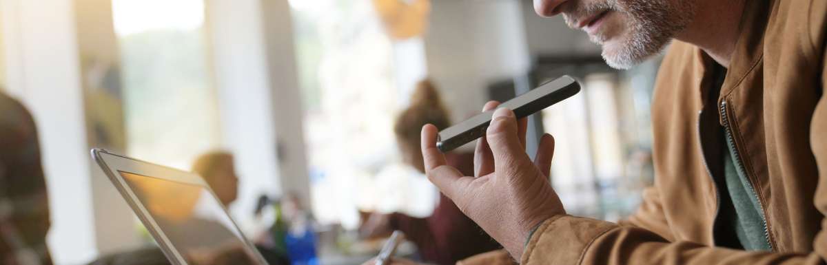 A middle-aged man sitting in a busy coffee shop and speaking into his cellphone while viewing his open laptop.