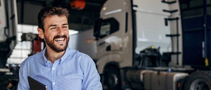 A worker wearing a blue shirt and holding a tablet smiles while two semi-trucks sit behind him in a large warehouse.