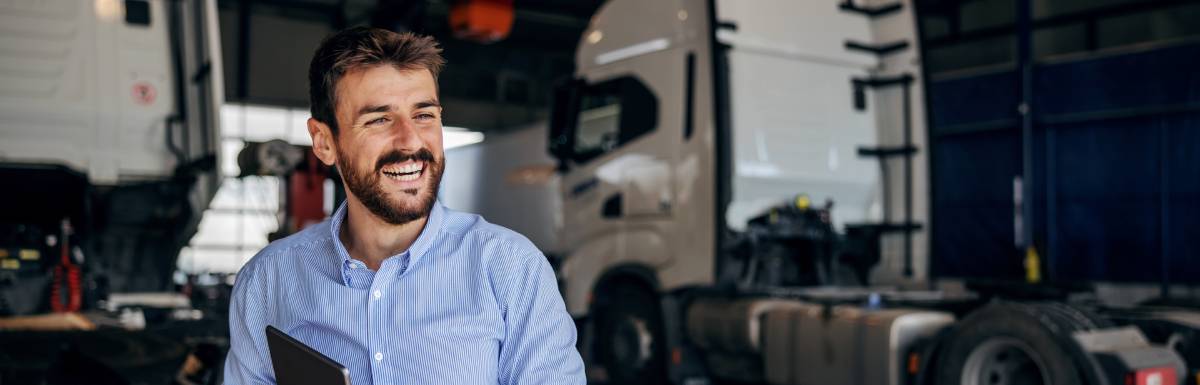 A worker wearing a blue shirt and holding a tablet smiles while two semi-trucks sit behind him in a large warehouse.