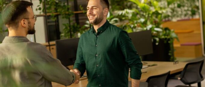 Two male business colleagues shaking hands in the workplace. There are plants and living elements in the office area.