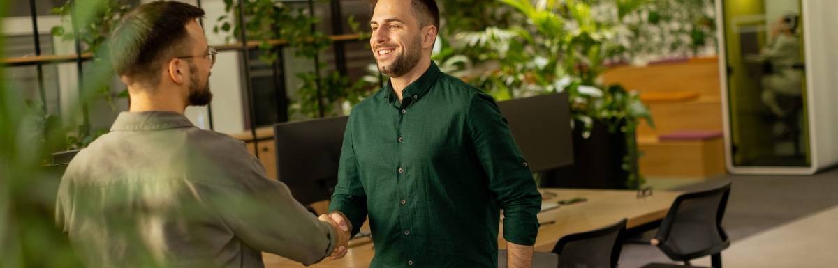 Two male business colleagues shaking hands in the workplace. There are plants and living elements in the office area.