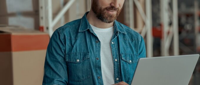 A man is standing inside a warehouse, holding an open laptop in his hand, and he works on it using his other hand.