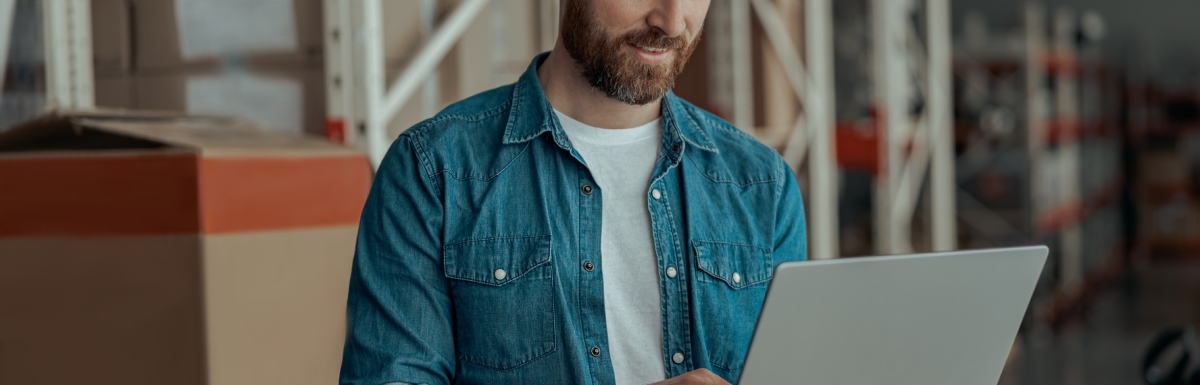 A man is standing inside a warehouse, holding an open laptop in his hand, and he works on it using his other hand.