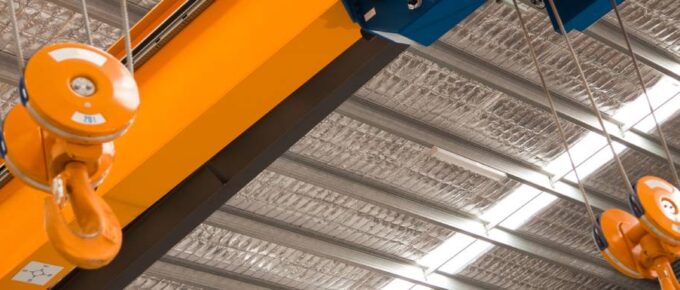 A blue and yellow overhead crane suspended from the ceiling of an industrial facility with large steel beams and recessed lighting.