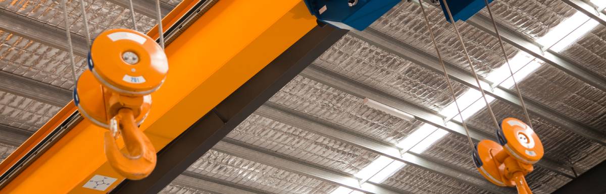 A blue and yellow overhead crane suspended from the ceiling of an industrial facility with large steel beams and recessed lighting.