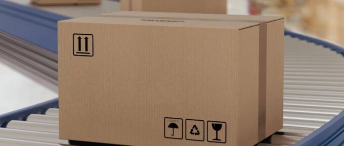 A cardboard box moves on a conveyor roller system in a warehouse. Several other cardboard boxes are behind it.