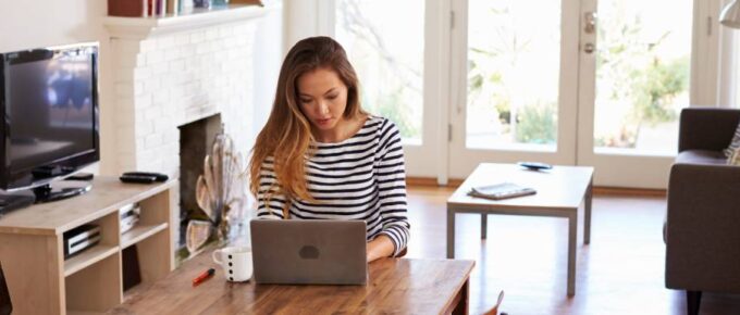 A woman in a striped shirt sitting at a wood table in front of a couch, coffee table, and TV using a laptop.