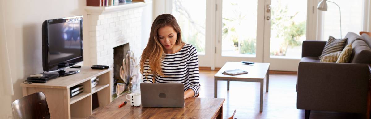 A woman in a striped shirt sitting at a wood table in front of a couch, coffee table, and TV using a laptop.