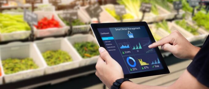 A man holds a tablet that reads "Smart Retail Management" while standing in the produce section of a grocery store.