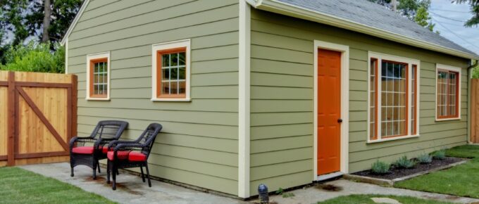 A green guest house with an orange door and orange paint trim. The house is in a fenced yard and has a small concrete patio.