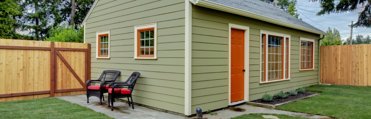 A green guest house with an orange door and orange paint trim. The house is in a fenced yard and has a small concrete patio.