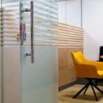 A space in an office separated by glass doors. The space has a yellow cushioned chair and a table.