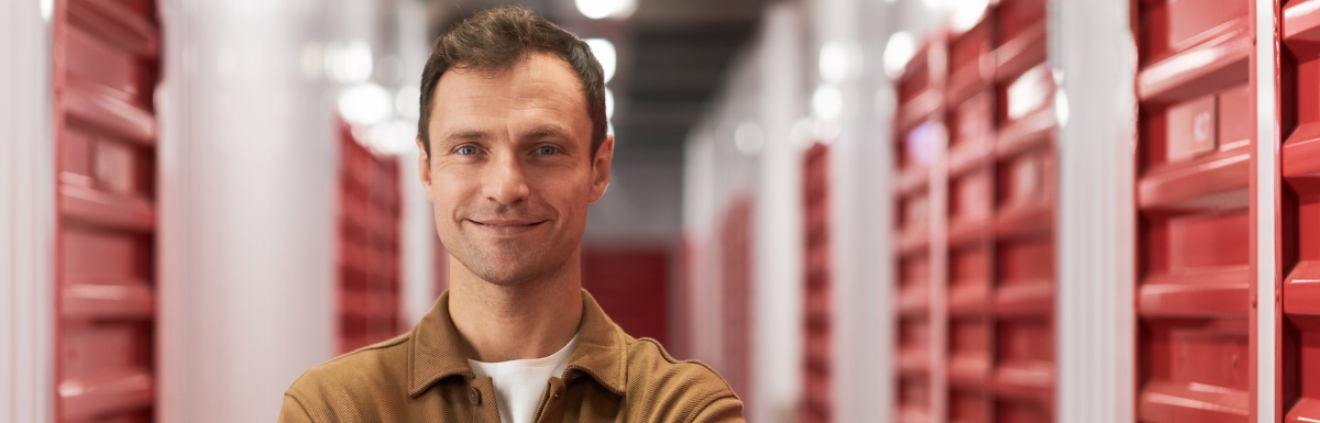 A man stands in front of storage units with red doors, arms crossed and smiling confidently, appearing proud.